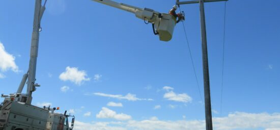 Outdoor photograph of a bucket truck lifting a worker within reach of a transmission tower