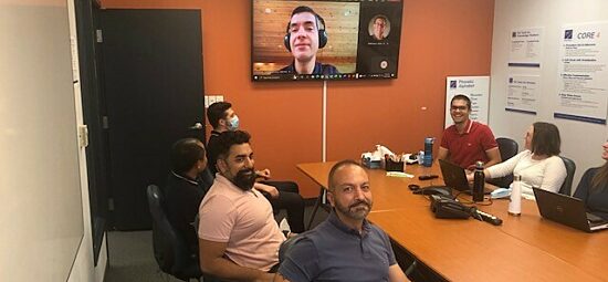 People meeting around a board table with a colleague joining virtually via screen on the wall.