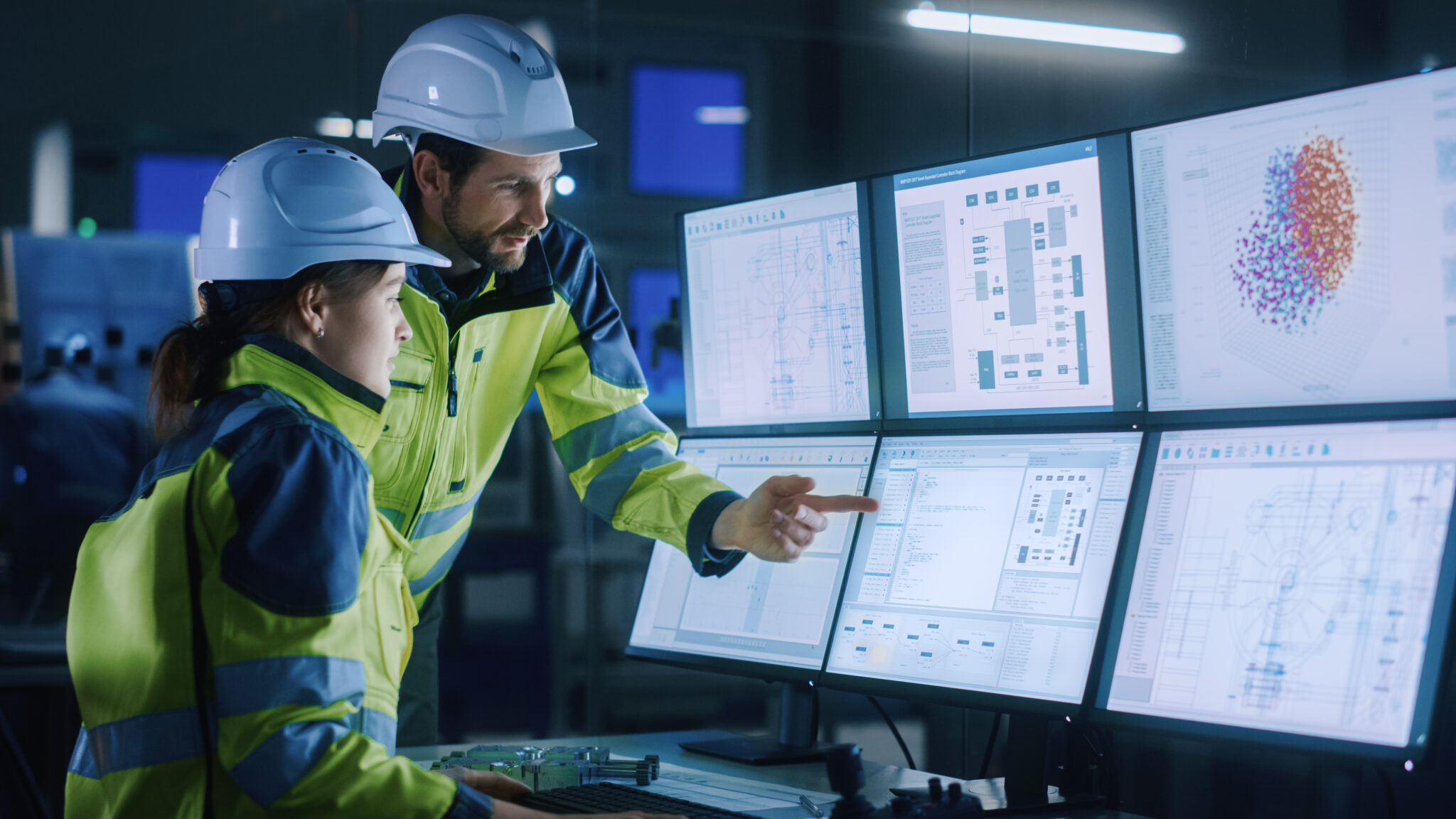 Engineers  in hard hats and hiviz jackets standing in front of a multi screened computer control system