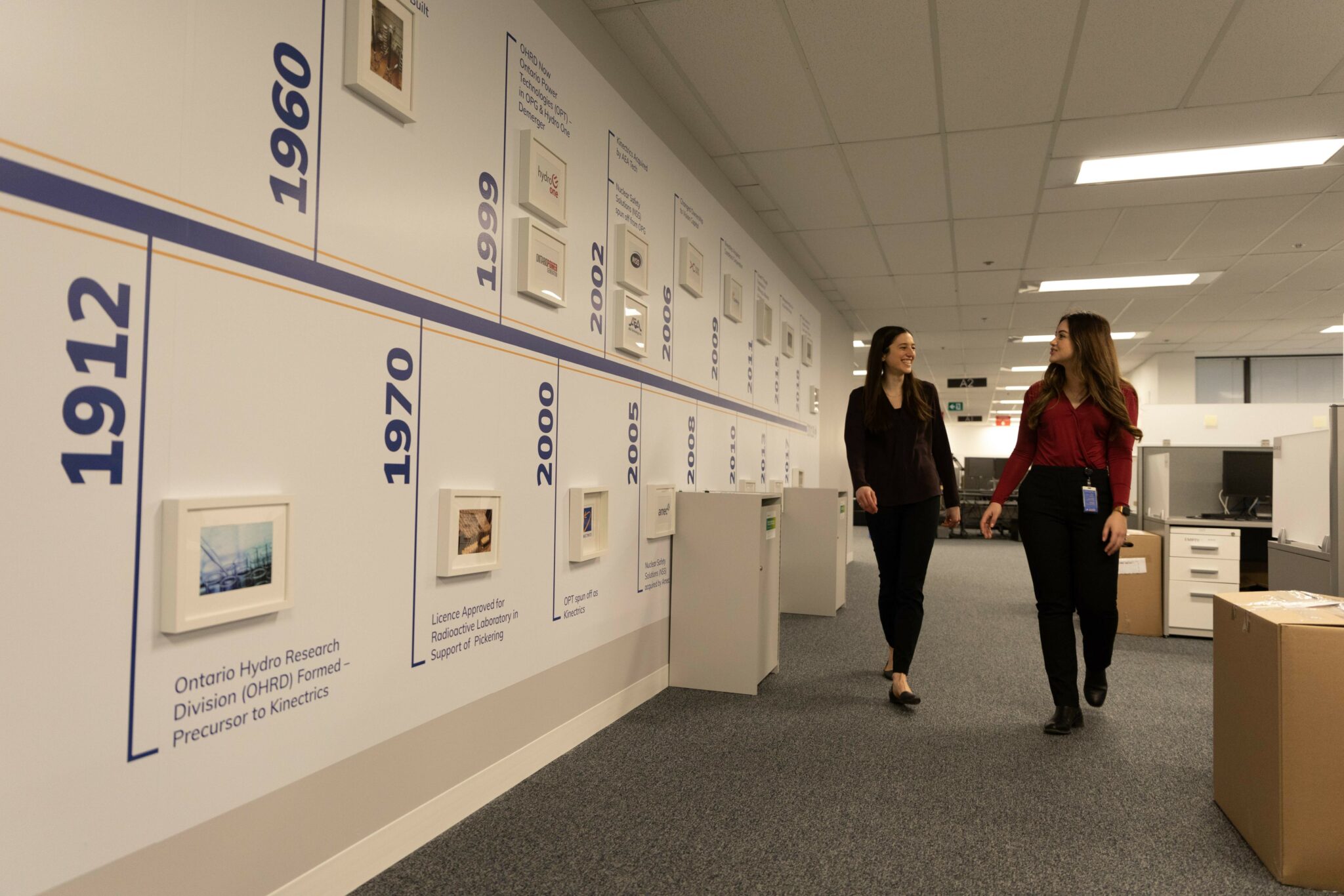 Two workers walking in front of a display wall showing the history of Kinectrics.