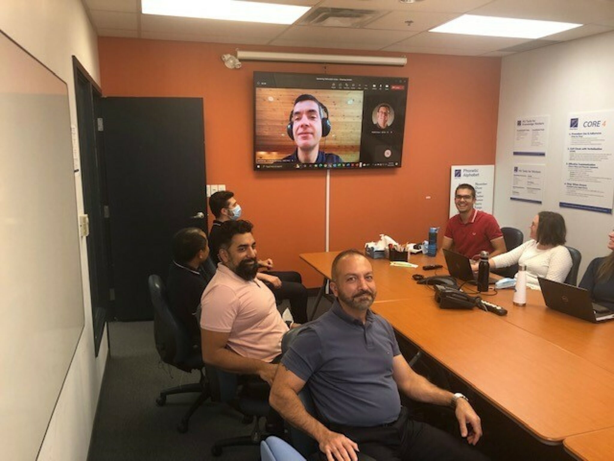People meeting around a board table with a colleague joining virtually via screen on the wall.
